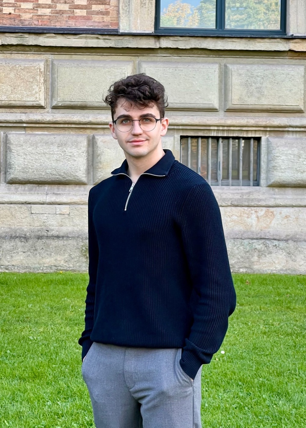 Nils Beham in a navy knit sweater and grey trousers, standing on a lawn in front of a sandstone building